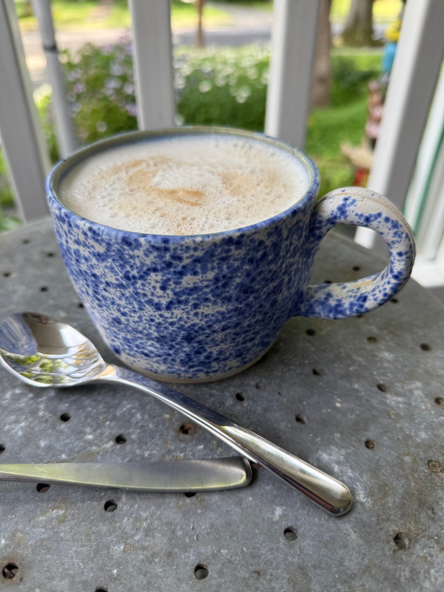 Close-up view of speckled blue glaze on handmade ceramic mug filled with cappuccino and surrounded by flatware in a casual outdoor setting